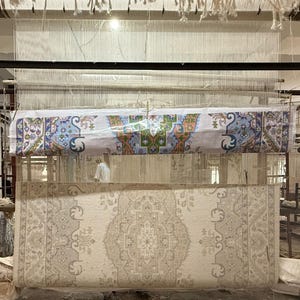 May include: A large loom featuring a partially woven rug with a beige, cream, and gray intricate floral pattern. Above the woven section, a printed paper guide displays a colorful design with blue, green, orange, and white geometric motifs.