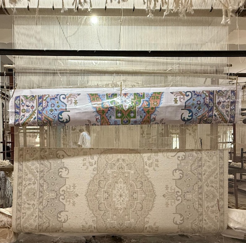 May include: A large loom featuring a partially woven rug with a beige, cream, and gray intricate floral pattern. Above the woven section, a printed paper guide displays a colorful design with blue, green, orange, and white geometric motifs.