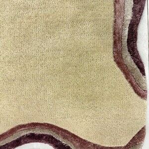 May include: A beige rug with a wavy, layered border in shades of burgundy, mauve, and taupe. The rug has a soft, textured surface and a unique, organic shape. The rug is designed for home decor.