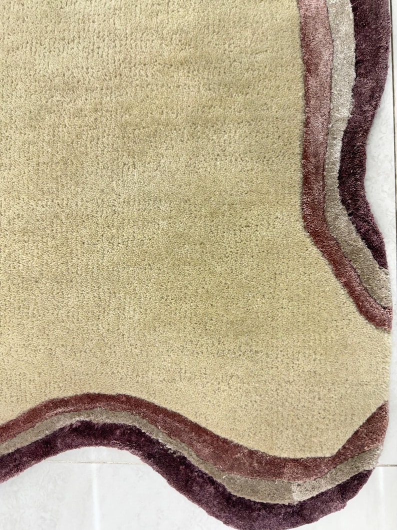 May include: A beige rug with a wavy, layered border in shades of burgundy, mauve, and taupe. The rug has a soft, textured surface and a unique, organic shape. The rug is designed for home decor.
