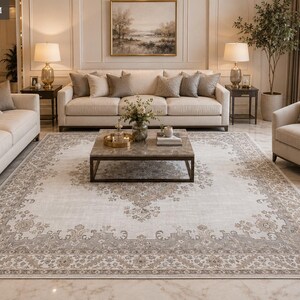 May include: A large rectangular area rug featuring a cream-colored center with a central medallion and a wide, ornate border in shades of beige and taupe. The rug displays a traditional floral pattern and is positioned in a living room setting.