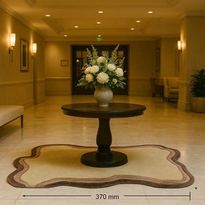 May include: A round, dark brown table with a vase of white flowers sits on a decorative rug in a well-lit hallway. The rug is approximately 14.5 inches wide. The hallway has a neutral color palette with sconce lighting.