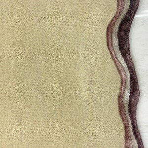 May include: A beige rug with a unique, organic shape. The rug features a textured surface and is bordered by layers of mauve, light gray, and dark purple. This decorative rug adds a touch of elegance to any room.