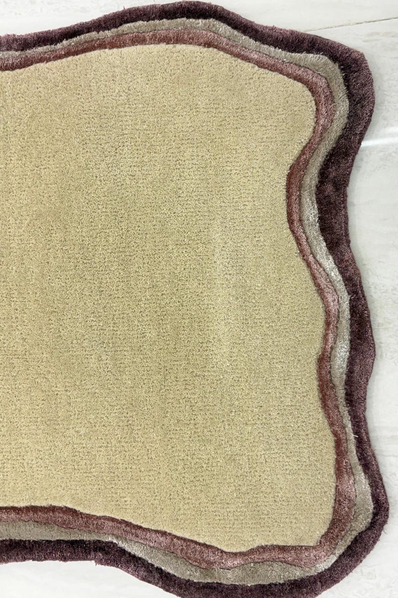 May include: A beige rug with a unique, organic shape. The rug features a textured surface and is bordered by layers of mauve, light gray, and dark purple. This decorative rug adds a touch of elegance to any room.