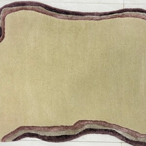 May include: A beige rug with an irregular, organic shape. The rug features a layered border in shades of brown and mauve, creating a textured, three-dimensional effect. The rug's design is modern and would complement a variety of interior styles.