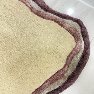 May include: A beige, wavy-edged rug with three layered borders in shades of mauve, light gray, and deep purple. The rug has a soft, textured surface and is likely made of a plush material. The design is modern and abstract.