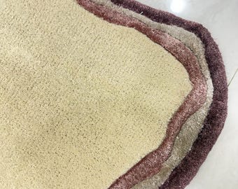 May include: A beige, wavy-edged rug with three layered borders in shades of mauve, light gray, and deep purple. The rug has a soft, textured surface and is likely made of a plush material. The design is modern and abstract.
