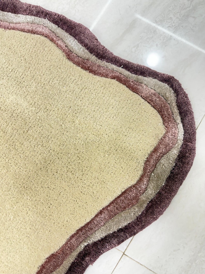 May include: A beige, wavy-edged rug with three layered borders in shades of mauve, light gray, and deep purple. The rug has a soft, textured surface and is likely made of a plush material. The design is modern and abstract.