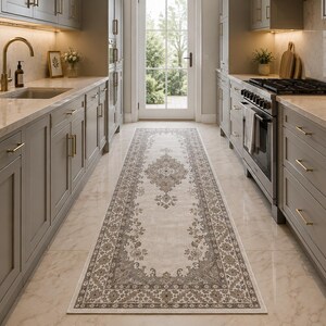 May include: A long, rectangular runner rug featuring a cream-colored center with a detailed, symmetrical gray floral medallion pattern and a matching ornate border, positioned on a light-colored floor between two rows of gray kitchen cabinets.