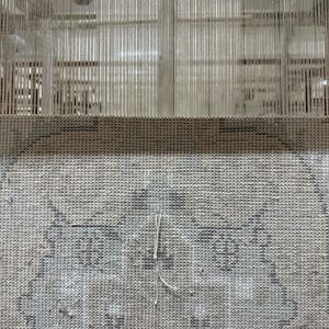May include: A woven textile piece featuring a light gray and white intricate pattern with a visible grid structure. The fabric is attached to a vertical loom with numerous thin, taut, light-colored warp threads extending upward from the top edge.