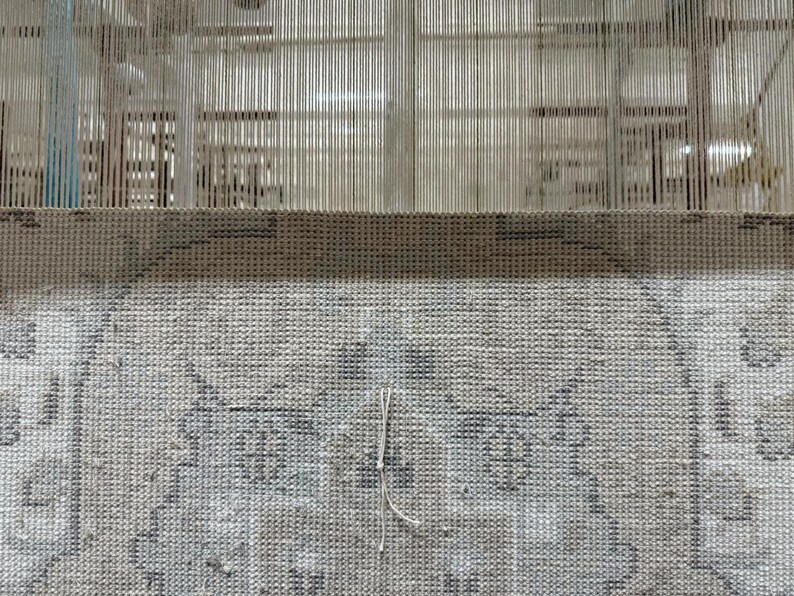 May include: A woven textile piece featuring a light gray and white intricate pattern with a visible grid structure. The fabric is attached to a vertical loom with numerous thin, taut, light-colored warp threads extending upward from the top edge.