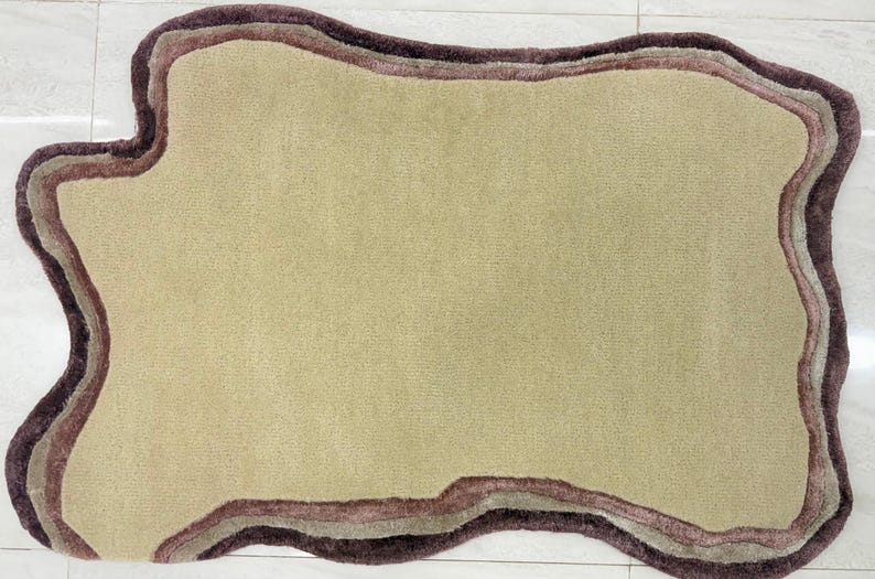 May include: A beige rug with an irregular, organic shape. The rug features a layered border in shades of brown and mauve, creating a textured, three-dimensional effect. The rug's design is modern and would complement various interior styles.