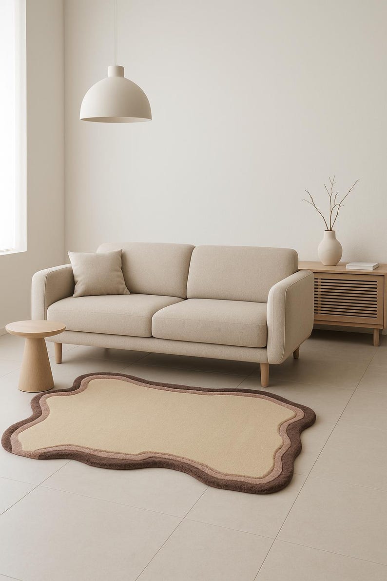 May include: A neutral-toned living room scene featuring a beige sofa, a small wooden side table, and a unique rug. The rug has an organic shape with brown and beige borders. A matching beige pillow rests on the sofa.