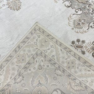 May include: A woven area rug featuring a light cream and beige color palette with intricate floral and geometric patterns. The design includes a detailed border and a central field with repeating motifs in shades of taupe, gray, and off-white.