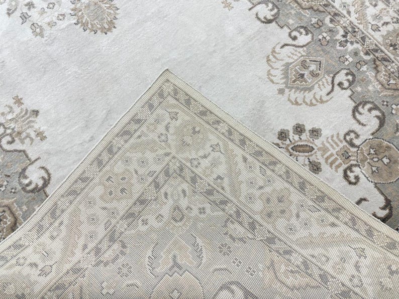 May include: A woven area rug featuring a light cream and beige color palette with intricate floral and geometric patterns. The design includes a detailed border and a central field with repeating motifs in shades of taupe, gray, and off-white.