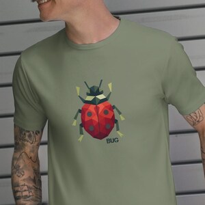 May include: Olive green t-shirt featuring a geometric ladybug design with a red body, black head, and the word "BUG" below. The ladybug has yellow and green accents. The shirt is short-sleeved.
