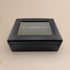 May include: A black square box with a clear top, featuring the gold text "GIVENCHY". The box has a sleek, minimalist design. The brand name is also visible on the bottom of the box.