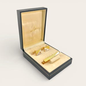 Puede incluir: Un juego de gemelos y un pasador de corbata dorados presentados en una caja negra con interior color crema. Los gemelos y el pasador de corbata presentan un diseño a rayas blancas y doradas. La caja tiene la marca "dunhill".