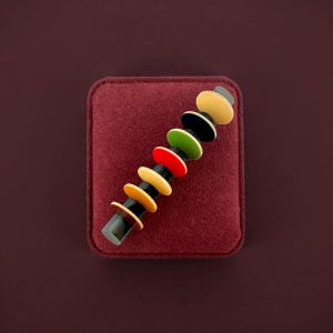 Peut inclure: Broche vintage avec une tige noire et des accents circulaires colorés en jaune, noir, vert, rouge et crème. La broche est présentée sur une surface en velours bordeaux, mettant en valeur son design rétro.