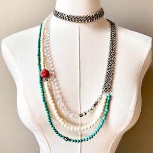 Könnte beinhalten: Halsketten-Set auf einer weißen Schaufensterpuppe. Das Set besteht aus einem silbernen Choker, einer langen Silberkette und Perlenketten in Türkis, transparent und creme. Eine rote Blumenperle und eine dekorative silberne Perle sind zu sehen.