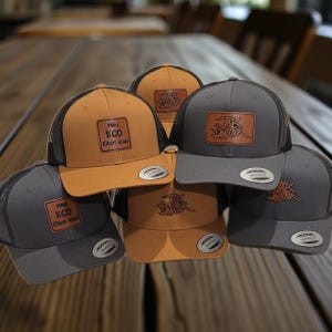 May include: A collection of trucker hats in shades of gray and tan, featuring mesh backs and leather patches. Some hats have the text "Make ECO Great Again" and others display the "JAK DAWGER" logo. The hats are arranged on a wooden surface.