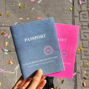 May include: Two rectangular booklets resembling passports, one in slate blue and one in bright pink. Both feature the text PASSPORT, A JO & SPENCER ADVENTURE, EURO BIKE TOUR EDITION, and a circular stamp with a bicycle icon reading NORWAY TO TURKEY.