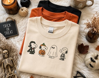 May include: A stack of three crew neck t-shirts in cream, orange, and black. The top cream shirt features a graphic of a cartoon dog dressed as a vampire, a pumpkin, a ghost, and a skeleton. The shirts are displayed on a white faux fur surface.