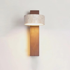 May include: A modern wall sconce with a cylindrical, speckled beige lampshade and a rectangular, wooden base. The sconce emits a warm, inviting light, perfect for interior decor. The fixture is mounted on a white wall.