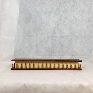 May include: A rectangular wooden display shelf with a dark brown top and a light wood base. The base is adorned with evenly spaced brass bullet casings, creating a unique and industrial aesthetic.