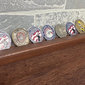 May include: A wooden display stand holding seven commemorative coins. The coins feature patriotic designs with red, white, and blue colors, and various military-themed imagery. The stand is made of dark wood and sits on a wooden surface.