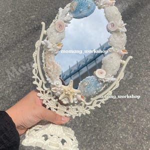 Könnte beinhalten: Ein ovaler Schminkspiegel mit einem weißen, verzierten Rahmen. Der Rahmen ist mit Muscheln, Perlen und einem Seestern verziert. Der Spiegel hat einen weißen Sockel und wird von einer Hand gehalten.