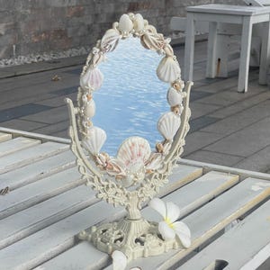 Peut inclure: Un miroir de courtoisie ovale avec un cadre crème orné de coquillages et de perles. Le miroir est posé sur un support décoratif et repose sur une surface en bois blanc. Des fleurs blanches sont éparpillées autour de la base du miroir.