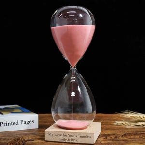 Puede incluir: Un reloj de arena de cristal transparente con arena rosa, sobre una base de madera. La base tiene grabado el texto "My Love for You is Timeless. Emily & David." Este reloj de arena es un elemento decorativo.
