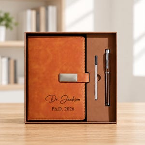 Pode incluir: Um caderno de couro sintético castanho com um fecho magnético prateado e o texto gravado Dr. Jackson Ph.D. 2026, apresentado numa caixa de presente com uma caneta de metal a condizer e uma recarga de tinta, tudo num conjunto estruturado.