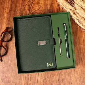Peut inclure: Un ensemble de journal et de stylo vert dans une boîte cadeau. Le journal a une couverture texturée avec un fermoir argenté et les initiales "MJ" en or. Un stylo vert assorti et une recharge de stylo sont inclus. L'ensemble est présenté sur une surface en bois.
