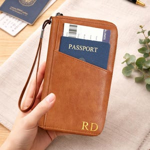 May include: A brown leather passport wallet with a zipper closure and a wrist strap. The wallet has a pocket holding a blue passport and a boarding pass. Gold initials "RD" are visible on the front.