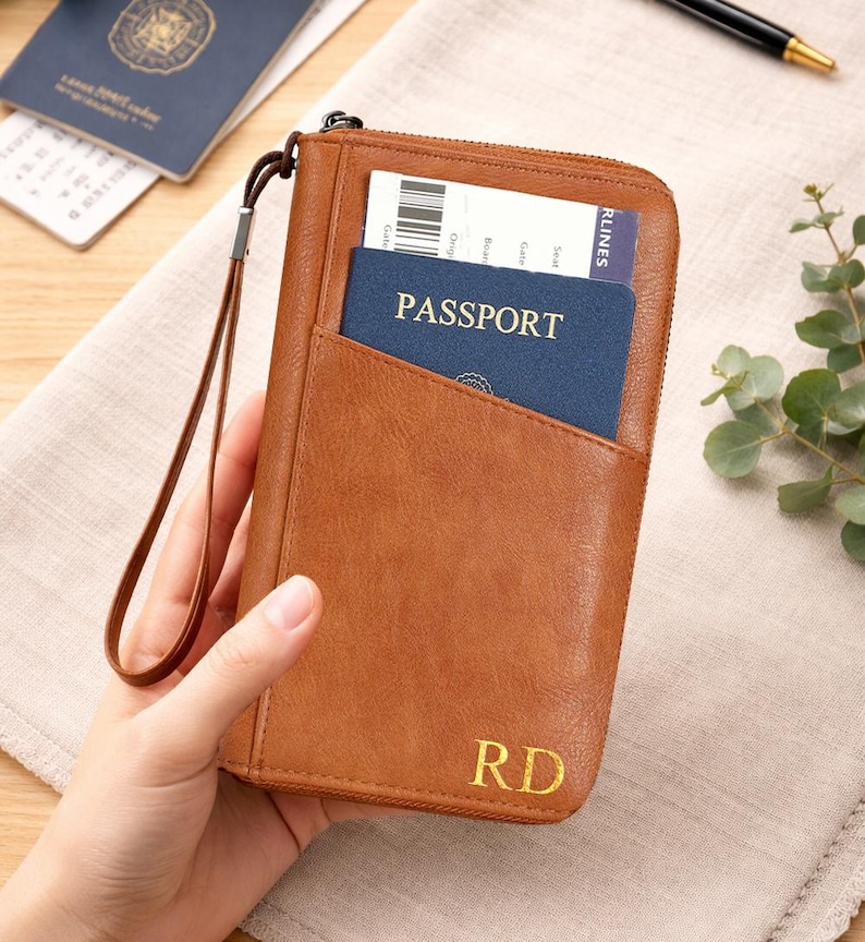 May include: A brown leather passport wallet with a zipper closure and a wrist strap. The wallet has a pocket holding a blue passport and a boarding pass. Gold initials "RD" are visible on the front.