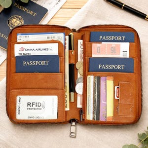 May include: Open brown leather travel wallet with multiple compartments holding passports, tickets, and currency. The wallet has a zip closure and RFID protection. Various cards and a pen are also visible.