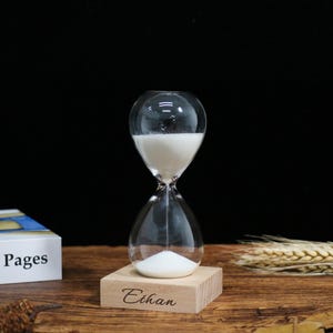 Könnte beinhalten: Eine transparente Glas-Sanduhr mit weißem Sand, die auf einem hellen Holzsockel steht. Der Sockel ist mit dem Namen "Ethan" graviert. Die Sanduhr steht vor einem dunklen Hintergrund, mit einem Buch und Weizenähren.
