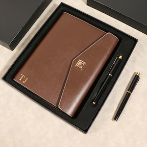 May include: A brown leather journal and pen set in a black gift box. The journal features the initials "TJ" in gold. Two black pens with gold detailing are included. The set is displayed on a light-coloured surface.