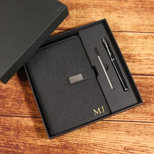 Puede incluir: Un juego de regalo negro con un cuaderno con cierre plateado y las iniciales "MJ" en dorado, junto con un bolígrafo y un lápiz óptico. El conjunto se presenta en una caja negra, ideal para regalar.