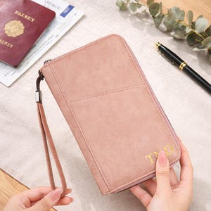 May include: A dusty rose travel wallet with a wrist strap and a zipper closure. The wallet has a textured surface and gold-colored initials. A passport and pen are visible in the background.