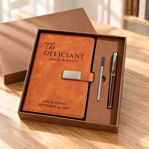 May include: A tan notebook with a silver metal clasp, featuring the text The OFFICIANT JAMES BENNETT and LIAM & SOPHIE SEPTEMBER 20, 2026, presented in a brown gift box alongside a dark brown pen and a silver ink refill cartridge.