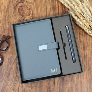 May include: A gray notebook and pen set in a gift box. The notebook has a tab closure and gold initials "MJ". The pen is silver and gray. The set is presented in a gray box on a wooden surface.