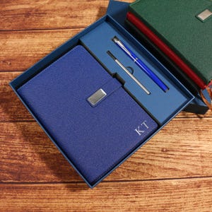 May include: A blue leather journal with a silver clasp and the initials "KT" is presented in a blue gift box. A blue pen and a silver pen are also included. A green journal is visible in the background.