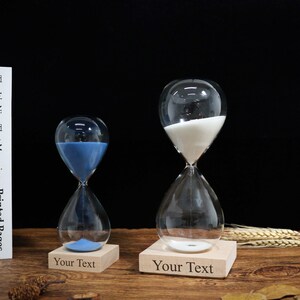Pode incluir: Duas ampulhetas de vidro transparente, uma com areia azul e outra com areia branca, assentam em bases de madeira. As bases estão gravadas com o texto "Your Text". As ampulhetas estão sobre uma superfície de madeira com fundo preto.