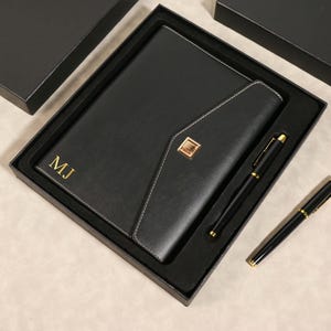 Op de afbeelding: Een zwart leren notitieboek en pennenset in een zwarte doos. Het notitieboek heeft de initialen "MJ" in gouden letters. De pen en het notitieboek hebben gouden accenten. Het notitieboek heeft een flap met een gouden vierkante sluiting.
