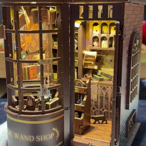 Puede incluir: Una maqueta de madera en miniatura de una tienda de varitas mágicas, con detalles interiores y exteriores. El letrero de la tienda dice "MAGIC WAND SHOP" y "Makers of Fine Wands since 1286". El modelo incluye libros, estanterías y una figura de gato.