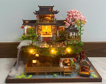 May include: Miniature Asian-style house with multiple levels, featuring a pagoda roof, red lanterns, and a small bridge. The scene includes a waterfall, a water wheel, and a pink flowering tree, all illuminated with warm lights.