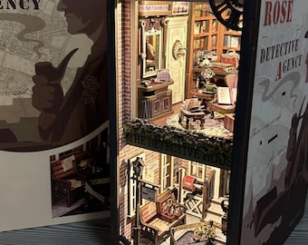 Może przedstawiać: Miniaturowa diorama agencji detektywistycznej z oświetleniem wewnętrznym. Model przedstawia dwupiętrowy budynek ze szczegółowymi meblami, półkami na książki i sceną uliczną. Na zewnętrznej skrzynce znajduje się napis "Rose Detective Agency".